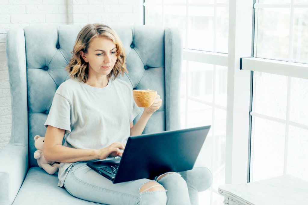 Young charming woman sits in a chair by the window with a cup of coffee and a laptop.